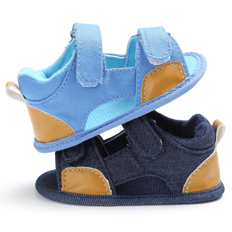 Two baby sandals stacked, one light blue and one dark blue, both with tan accents and Velcro straps. The sandals have soft soles and a fabric loop on the heel.