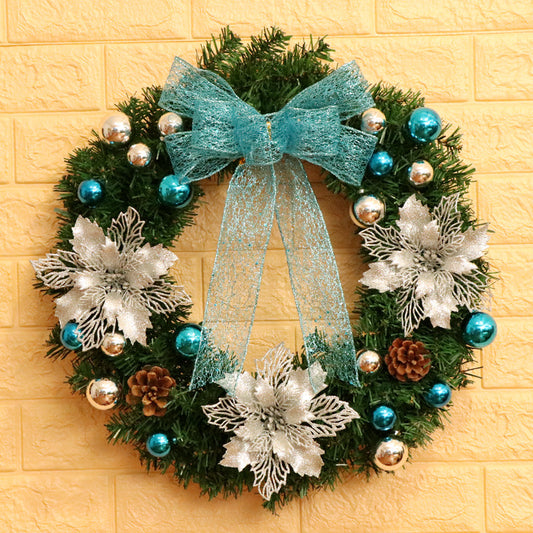 Festive wreath with a teal bow, silver flowers, blue and silver baubles, and pinecones on a green leafy base, hung on a yellow brick wall.
