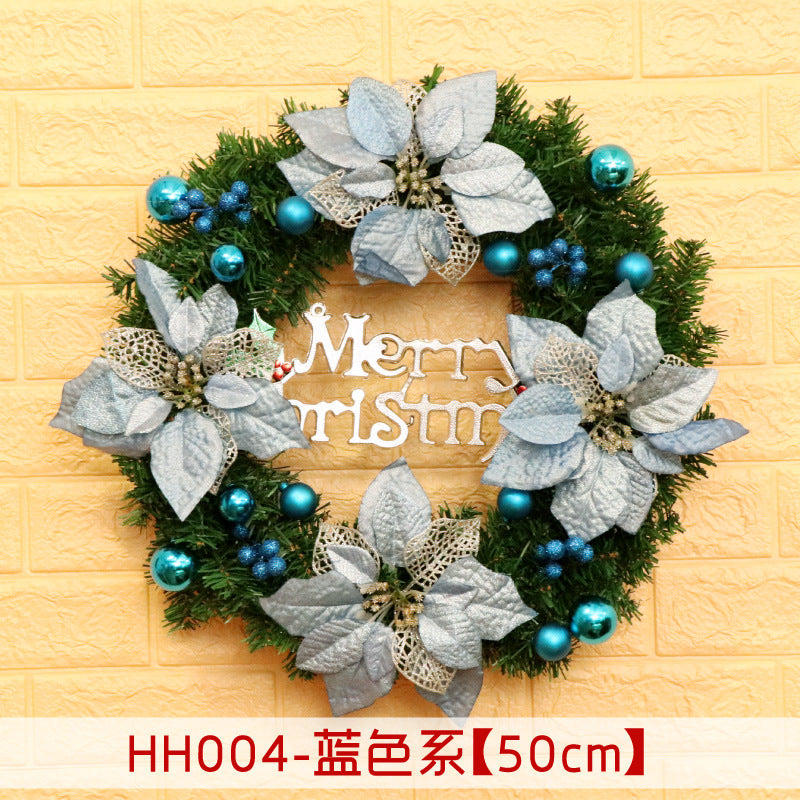 A green Christmas wreath featuring silver poinsettia flowers and blue ornaments, with a "Merry Christmas" sign in the center against a yellow textured wall.