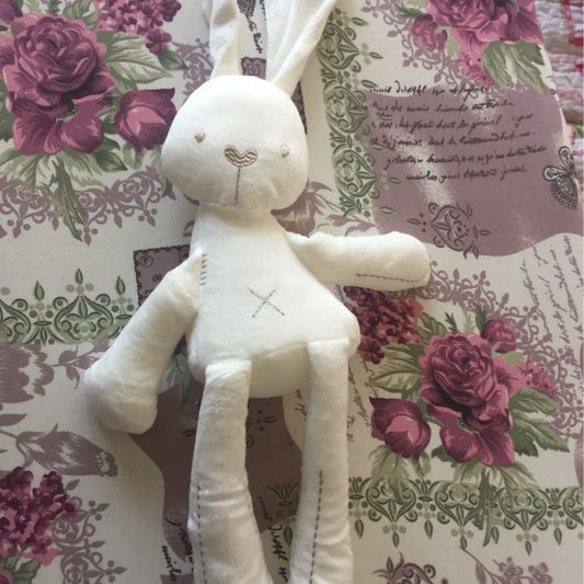 A soft, white stuffed rabbit toy with long ears and stitched facial features lies on a floral-patterned fabric adorned with pink roses and decorative script.