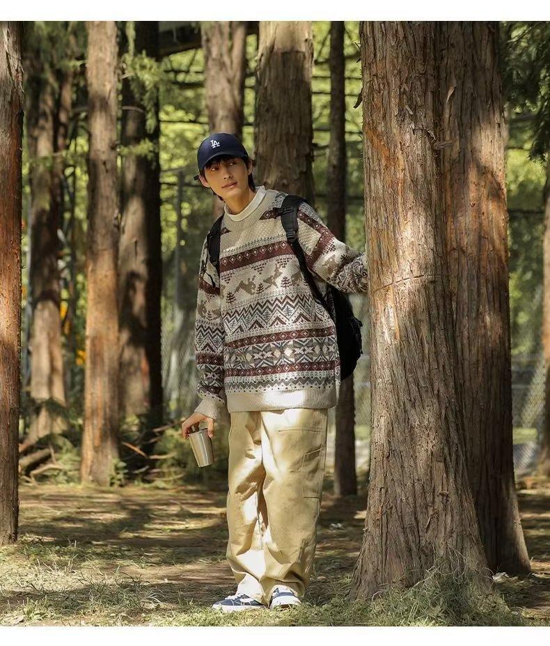 Person in a patterned sweater and beige pants stands by a tree in a sunlit forest, holding a silver mug. They wear a navy cap and backpack, surrounded by tall trees.