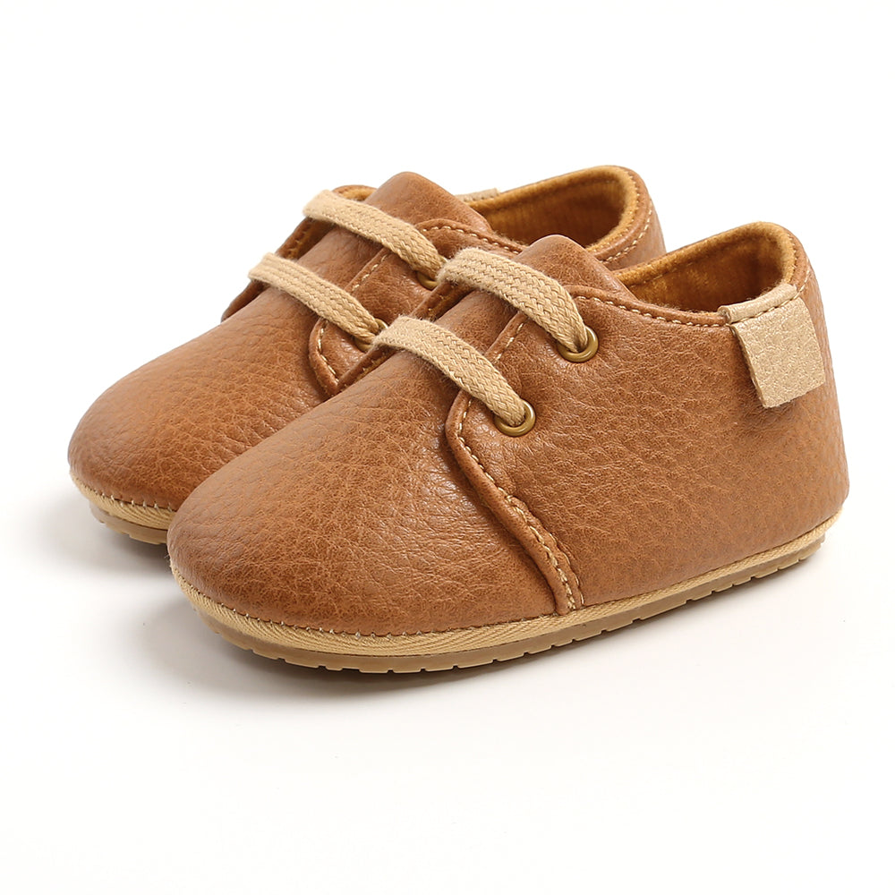 Brown baby shoes made of soft faux leather with beige laces and Velcro closure. The shoes have a textured appearance, and the soles are slightly ridged for grip.