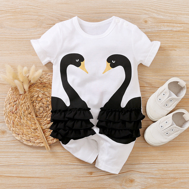 White baby romper with two black swans facing each other, adorned with ruffled black fabric. Set on a wooden surface, accompanied by white baby shoes and a woven mat with dried flowers.