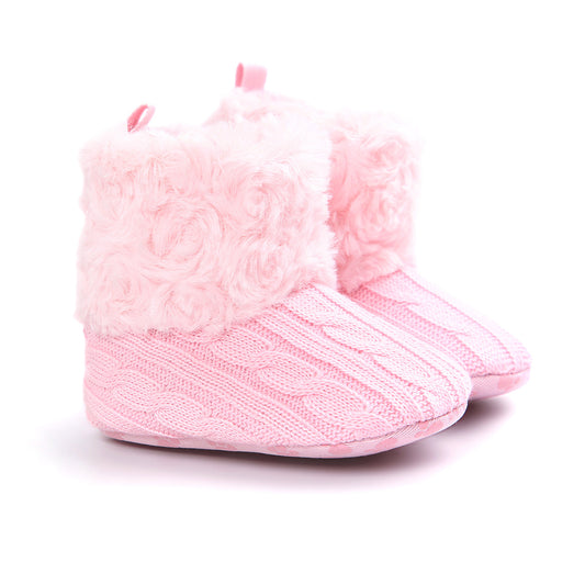 Pink baby booties with a knitted cable pattern and fluffy, textured cuffs. The booties have a soft, cozy appearance, ideal for keeping little feet warm.