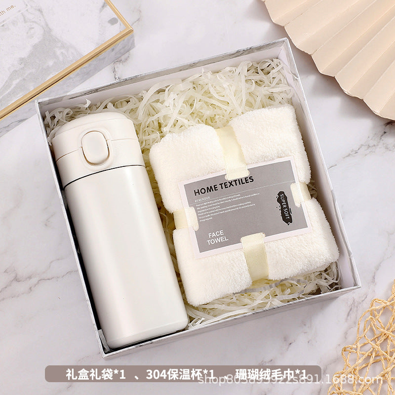 Gift box containing a white insulated travel mug and a soft, white face towel, nestled in shredded paper. Set on a marble surface with a decorative fan nearby.