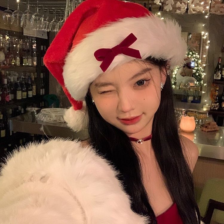 Young woman in a Santa hat winks at the camera in a cozy, festive setting with dim lighting. She has long dark hair and wears a red outfit, with holiday decorations in the background.