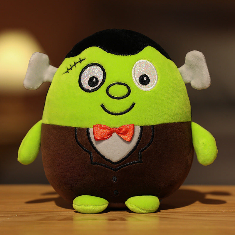 Cute plush toy resembling a cheerful green monster with a stitched smile, orange bow tie, brown suit, and playful bolts on its head, sitting on a wooden surface.