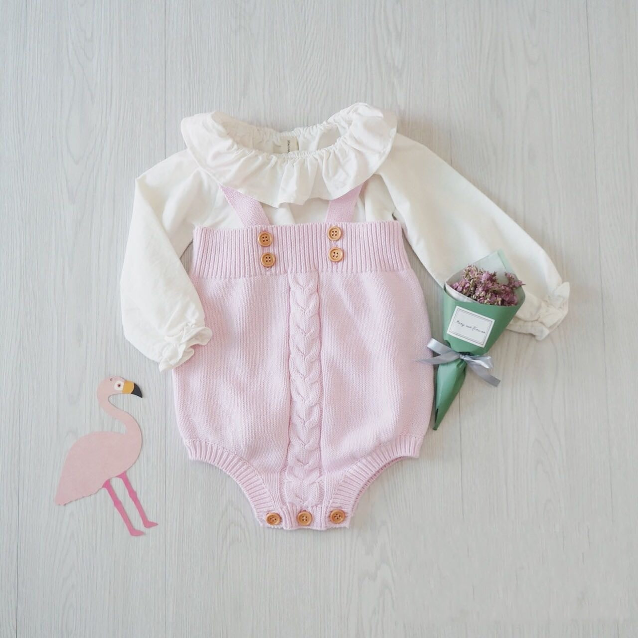 A baby outfit with a white ruffled top and pink knitted romper featuring buttons and a braided design, lies on a light wooden floor beside a pink flamingo cutout and small bouquet.