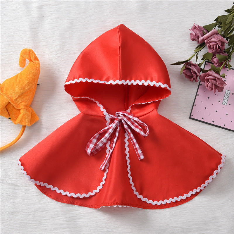 A red hooded cape with white scalloped trim and a gingham bow tie, laid on a white surface. An orange hat and pink roses in the background add color accents.