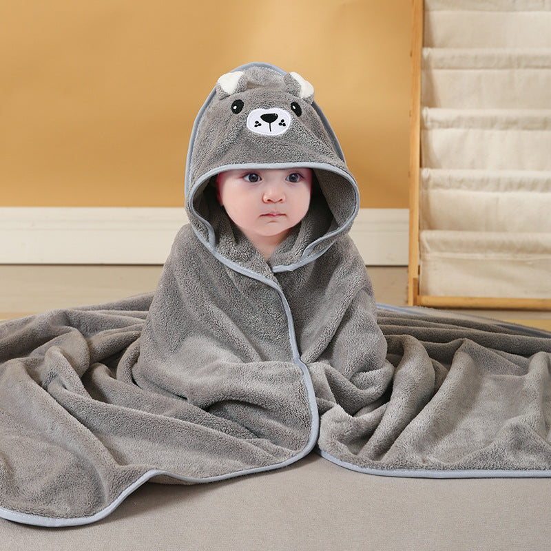 A baby wrapped in a soft, grey hooded towel with animal ears and a face, sitting on a beige rug against a light brown wall.