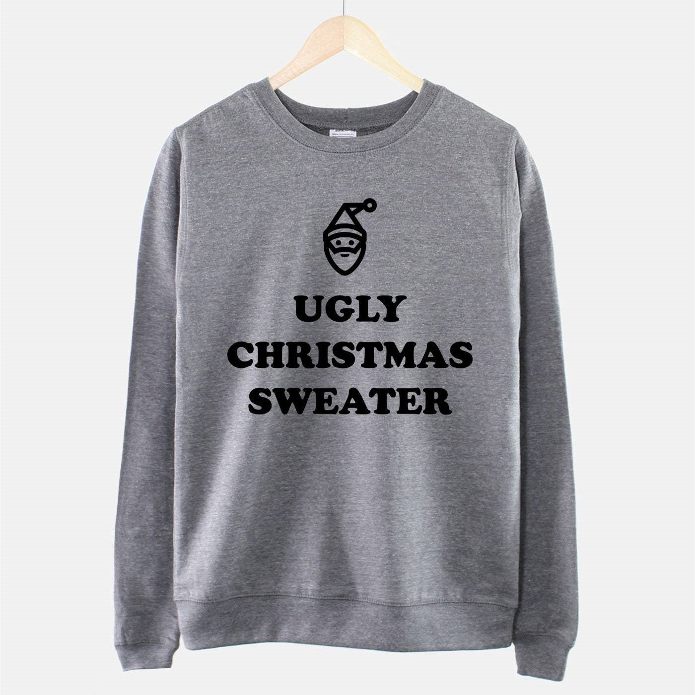 Gray sweatshirt hanging on a wooden hanger with the text "UGLY CHRISTMAS SWEATER" and a simple Santa face design in black on the front.