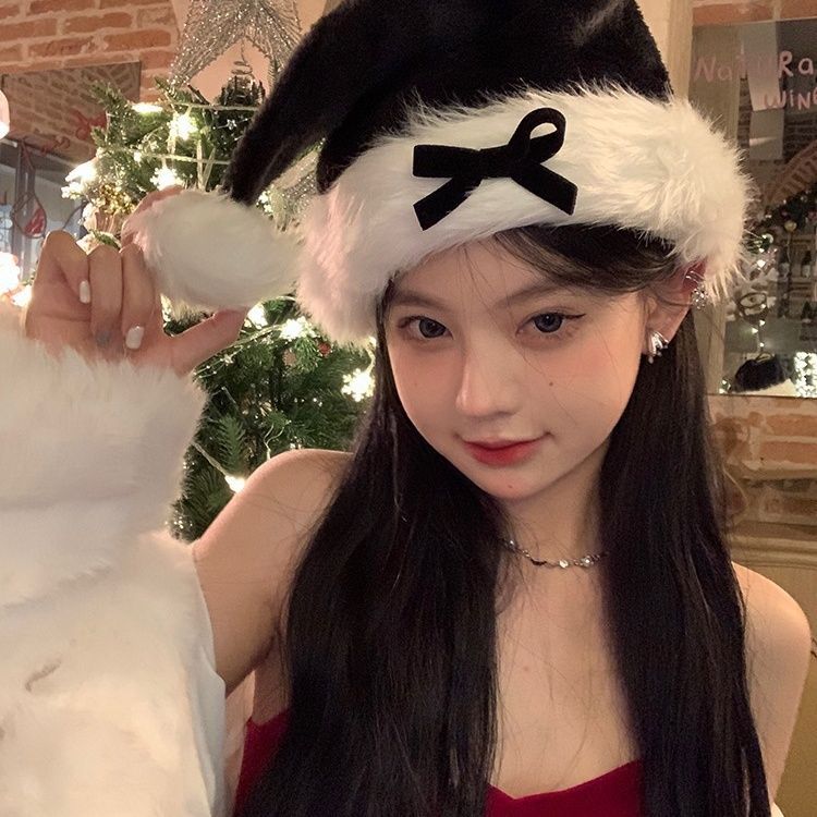 Young woman in a Santa hat with a black bow poses by a decorated Christmas tree. She holds the hat's pom-pom, wearing a red outfit and pearl necklace, smiling subtly.