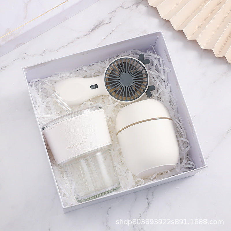Gift box with a white reusable tumbler, portable fan, and egg-shaped container, nestled in white shredded paper. Set on a marble surface beside a beige fan.