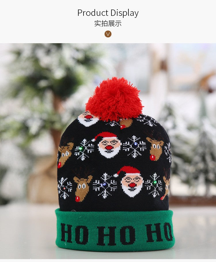 Festive knit hat with Santa and reindeer pattern, snowflakes, a red pom-pom, green cuff, and "Ho Ho Ho" text, displayed in a snowy background.