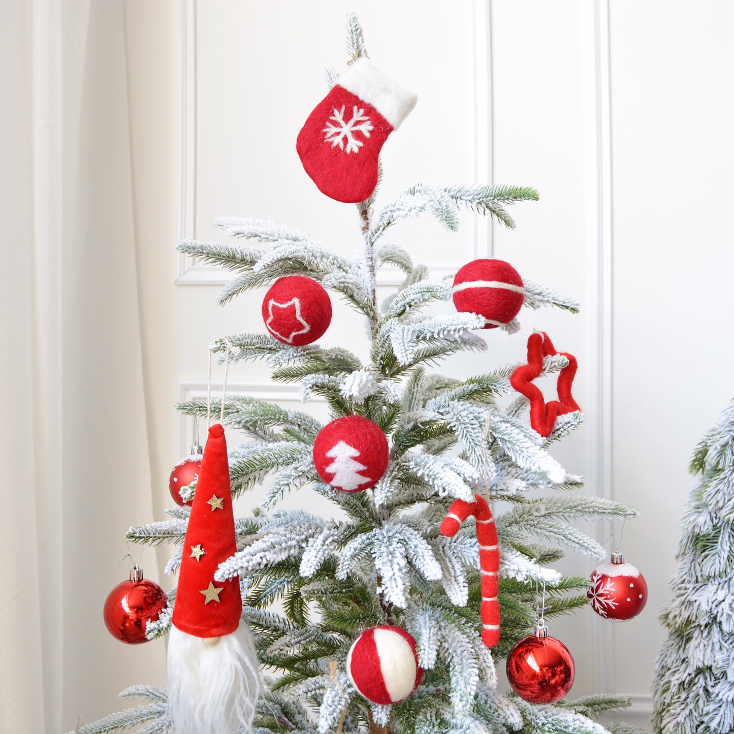 A small snow-covered Christmas tree adorned with red and white felt ornaments, including balls, a stocking, stars, a candy cane, and a Santa hat, against a white wall.