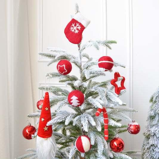 A small snow-covered Christmas tree adorned with red and white felt ornaments, including balls, a stocking, stars, a candy cane, and a Santa hat, against a white wall.