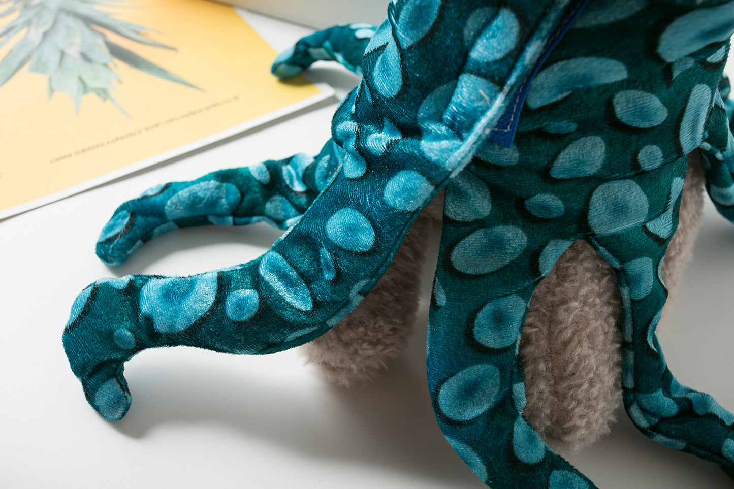 A plush octopus toy with teal and blue spotted legs, lies on a white surface near an open book with a leafy plant image on the visible page.