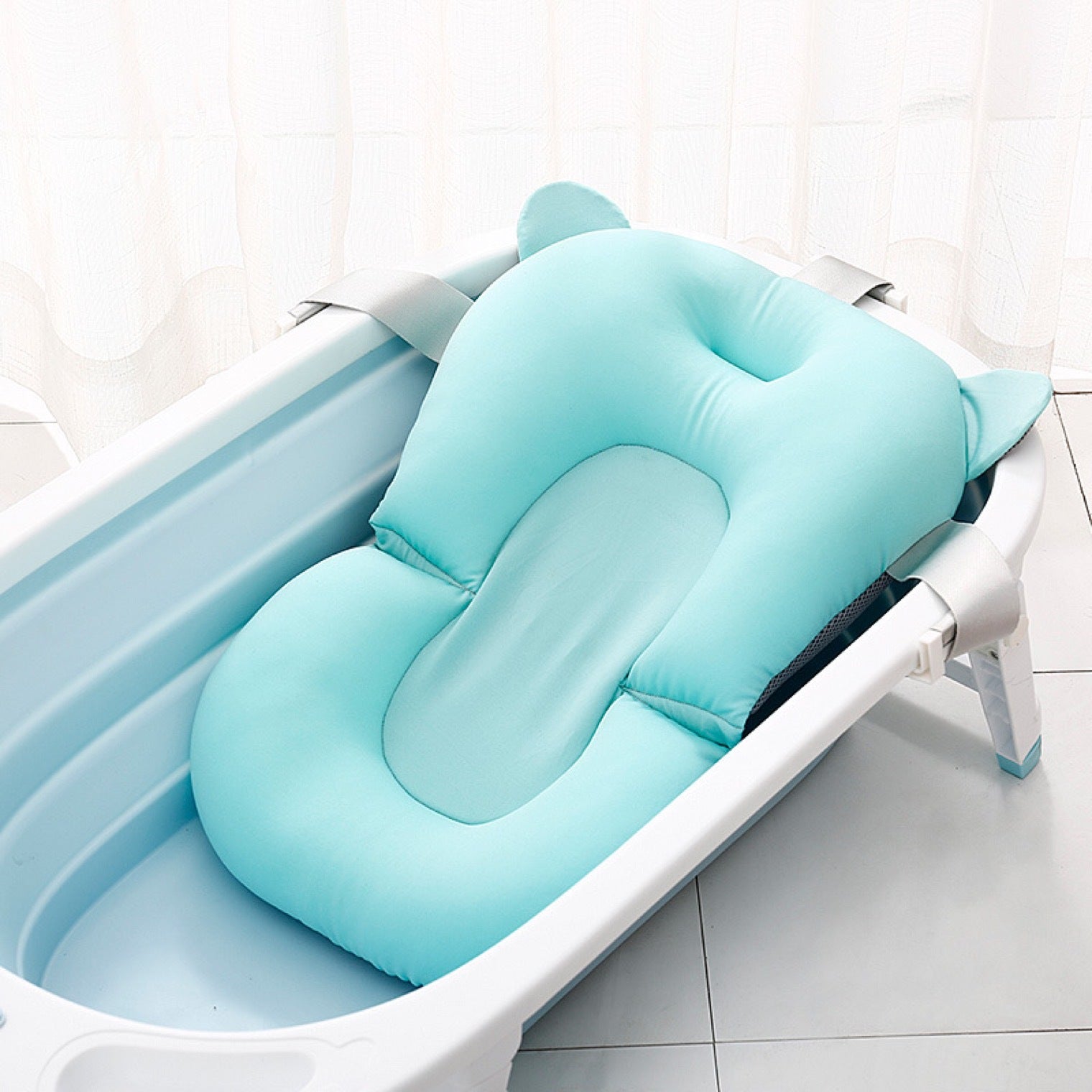 A light blue baby bathtub with a soft, cushioned insert featuring cute ear-like designs. The tub is placed on a tiled floor with white curtains in the background.