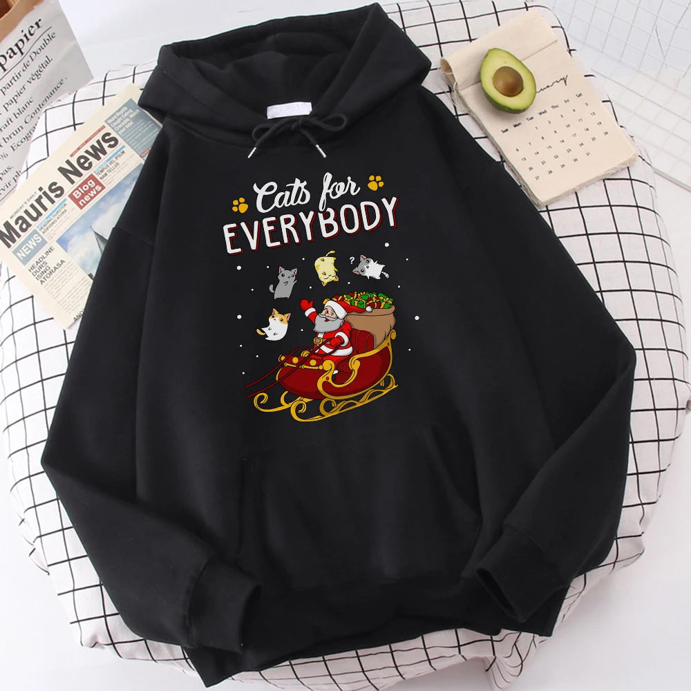 Black hoodie with "Cats for Everybody" text, featuring Santa in a sleigh surrounded by flying cats, on a checkered surface with a newspaper, a calendar, and half an avocado.