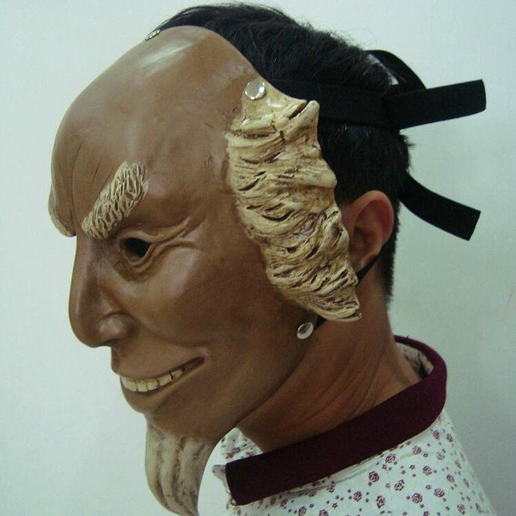 A person wearing a detailed, sculpted mask with exaggerated facial features, including pointed eyebrows, a thin beard, and large, textured ears. The mask is secured with black straps.