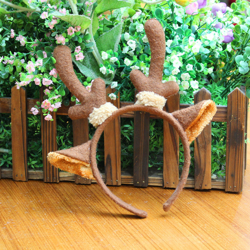 Brown felt headband with deer antlers and ears placed on a wooden surface. Background features a wooden fence and lush green plants with small pink and white flowers.