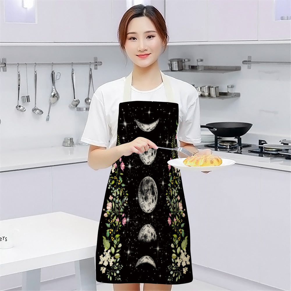 A woman in a kitchen wears a black apron with moon phases and floral patterns, holding a plate of pastry. Cooking utensils hang on the wall in the bright white kitchen.