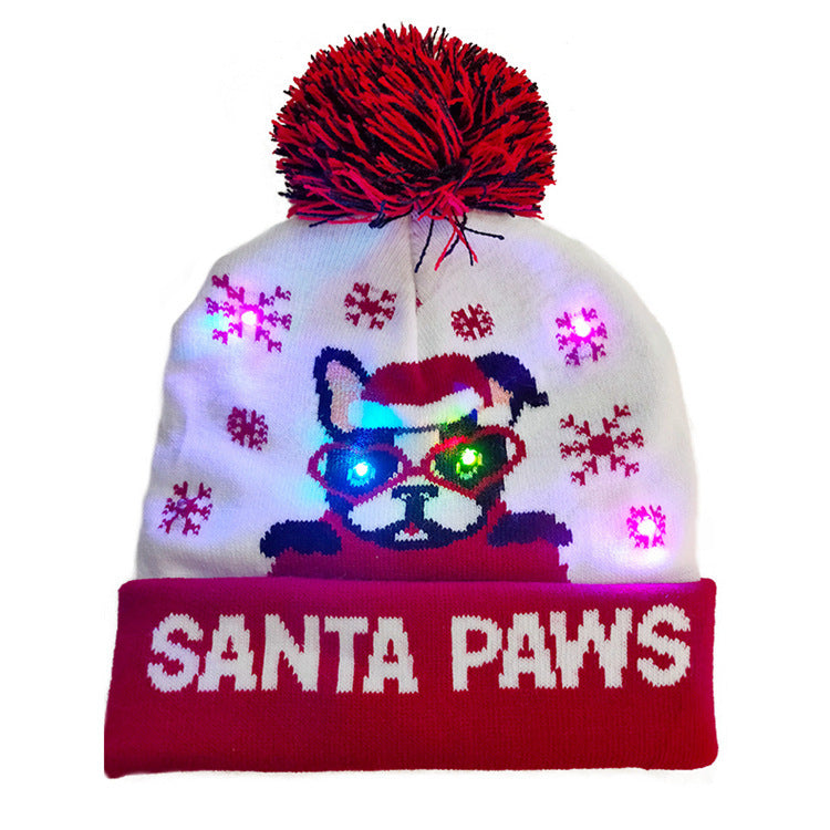 White beanie with a red pom-pom, featuring a cartoon dog in a Santa hat. LED lights illuminate the dog's eyes, surrounded by snowflakes. Red cuff reads "SANTA PAWS."