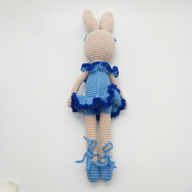 Crocheted bunny toy wearing a light blue dress with dark blue ruffles. The bunny has long ears, light pink limbs, and blue shoes with laces, lying on a white background.