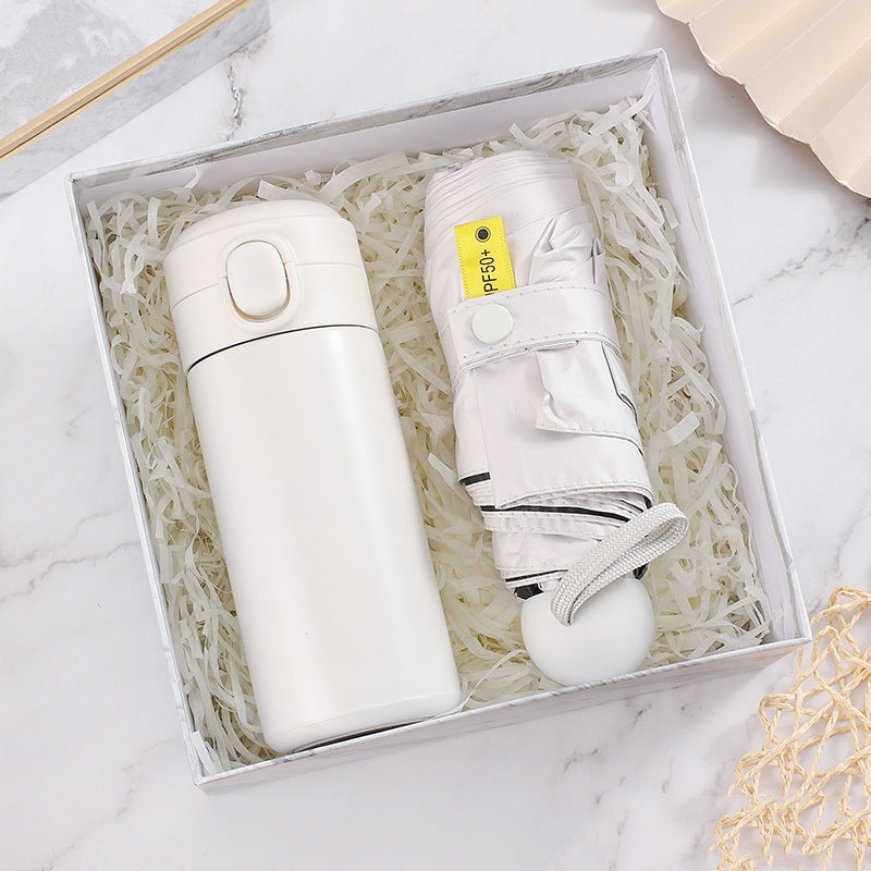 White travel mug and folded umbrella with "UPF 50+" tag, nestled in shredded paper inside a gift box. The items are placed on a marble surface.