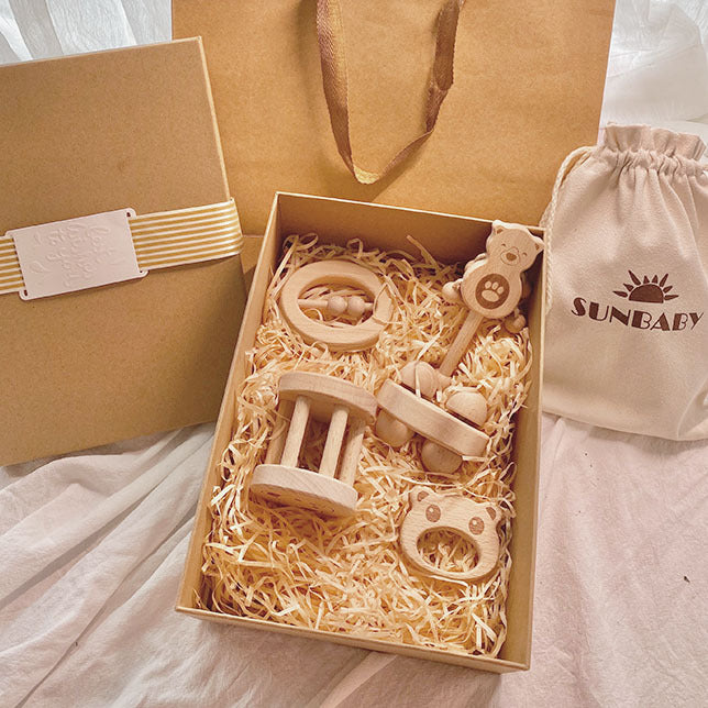 A gift box of wooden baby toys includes a rattle, teether, and beaded toys nestled in straw. A drawstring bag with a “SUNBABY” logo and closed cardboard box are beside it.