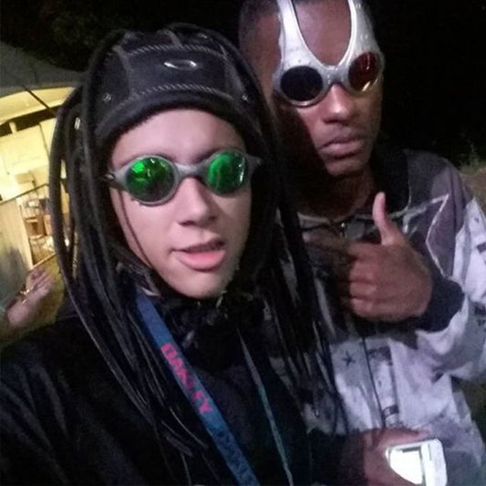 Two individuals wearing futuristic eyewear and outfits. One has green-tinted goggles and a headpiece with black tendrils, while the other has silver glasses and a patterned jacket.