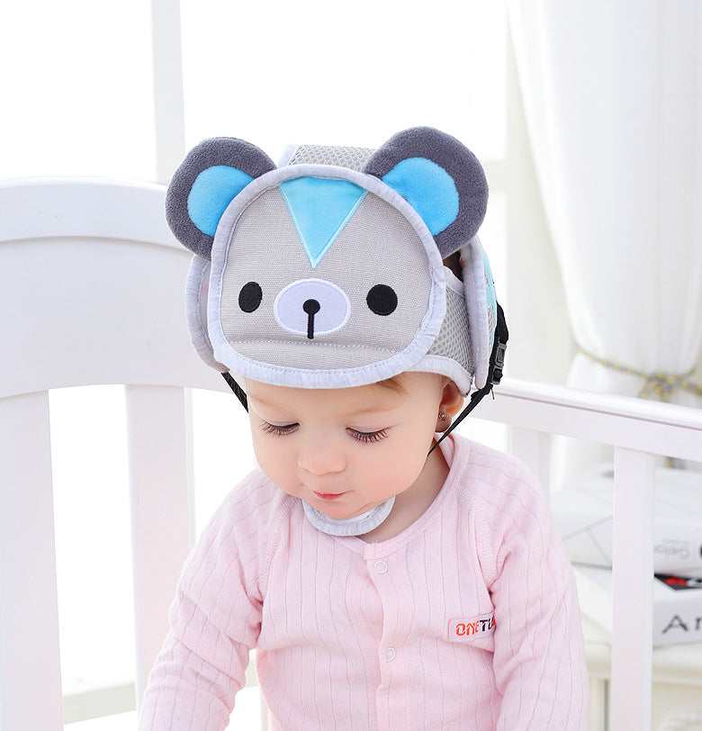 A baby wearing a pink onesie sits in a crib, wearing a gray and blue protective helmet with bear ears and face, against a bright background with white curtains.
