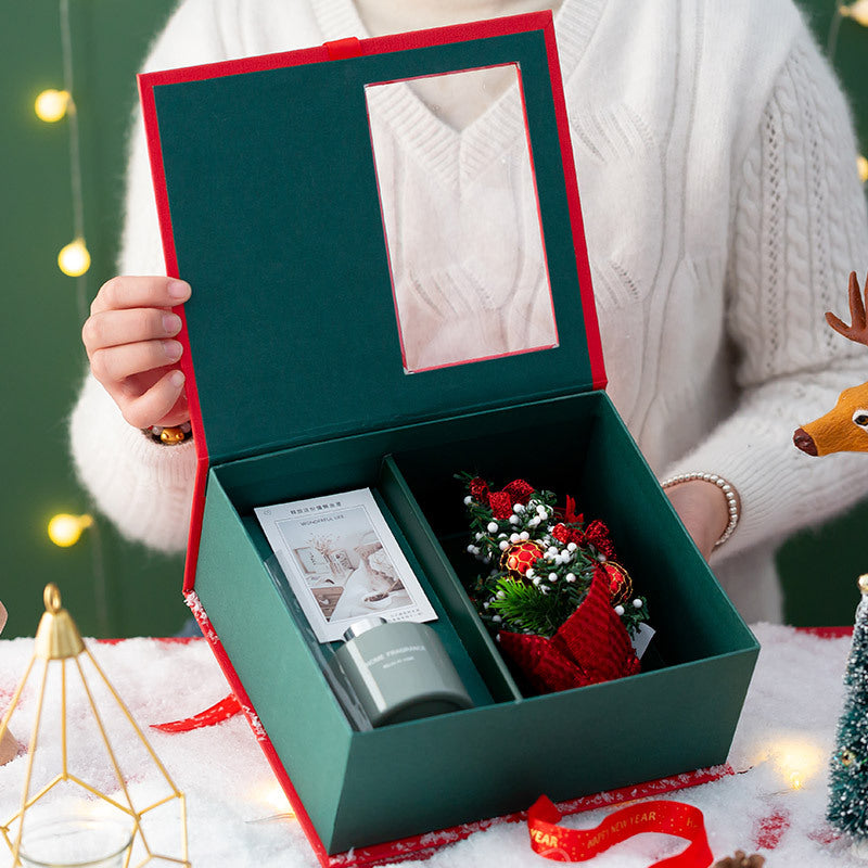 Person holding an open green gift box with red trim, revealing a small Christmas tree ornament and a candle inside. Warm fairy lights and festive decorations surround the scene.
