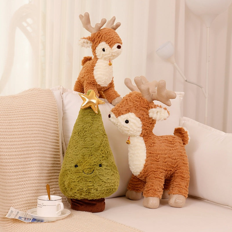 Two plush reindeer with bells stand beside a plush Christmas tree with a star on top. A cup and saucer with a spoon, and a magazine are on a beige sofa.