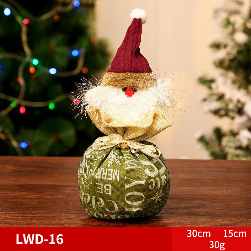 Festive gnome ornament with a red hat and fluffy beard, wrapped in green fabric with holiday text. It sits on a wooden table with a blurred Christmas tree in the background.