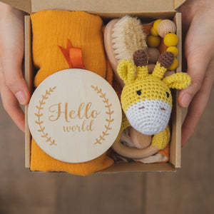 Hands hold a gift box with an orange blanket, wooden hairbrush, teether beads, a crocheted giraffe toy, and a round wooden sign reading "Hello world," adorned with a leafy border.