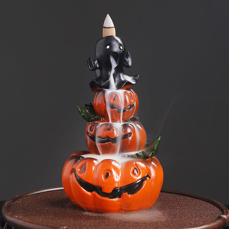 Ceramic incense burner with a ghost atop three stacked smiling pumpkins. Smoke flows downward, accentuating the Halloween theme, on a dark background.