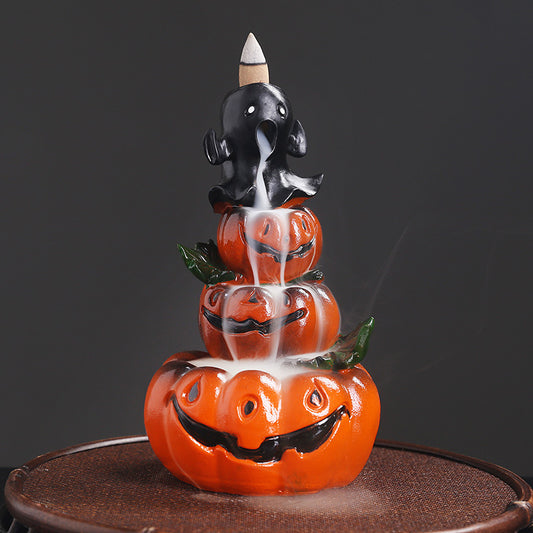 Ceramic incense burner with a ghost atop three stacked smiling pumpkins. Smoke flows downward, accentuating the Halloween theme, on a dark background.
