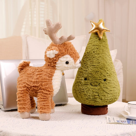 A plush reindeer toy stands beside a green plush Christmas tree with a gold star on top. Both rest on a white tablecloth with a laptop, cup, and newspaper in the background.