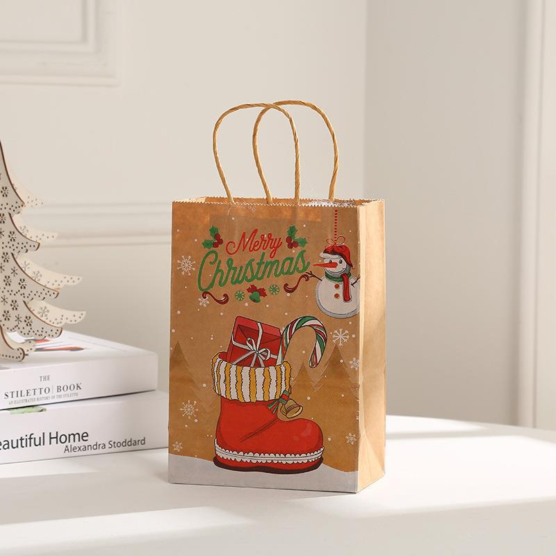 Brown gift bag with "Merry Christmas" text, red Santa boot with candy cane, snowflakes, and snowman design. It stands on a white surface near books and a decorative tree.