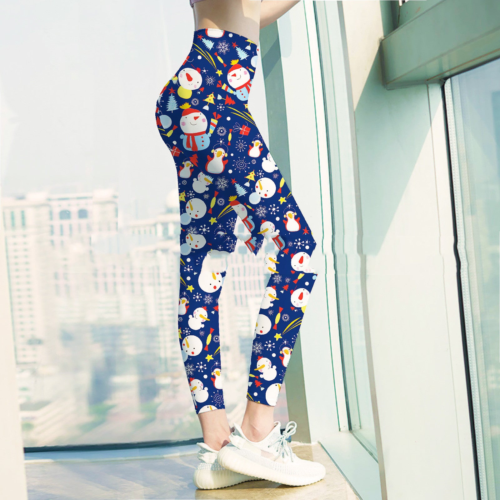 Person wearing festive blue leggings with snowmen and gifts, standing by a large window with a city view, paired with white sneakers.
