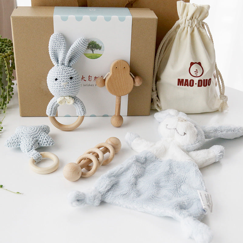 A collection of baby toys including a crocheted bunny rattle, wooden teether, and soft blue comforter, are arranged on a table with a gift box and drawstring bag.