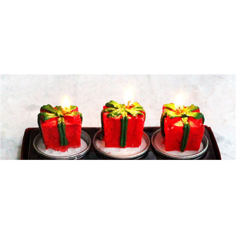 Three red candles shaped like gift boxes with green ribbons, each lit and placed on a small tray. The candles glow warmly against a light background.