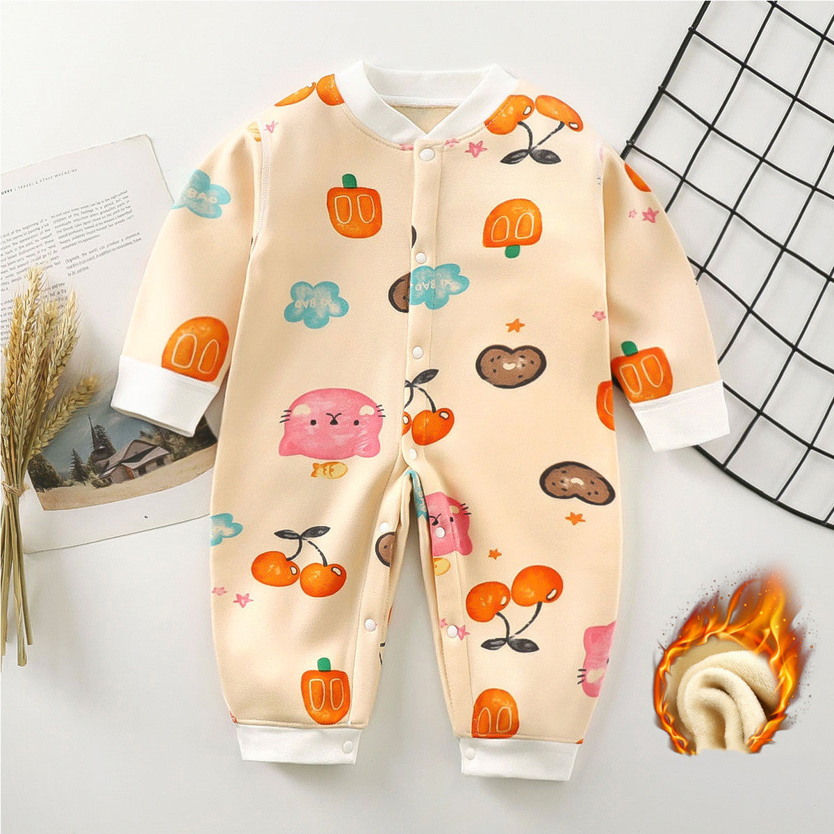 Beige baby onesie with colorful cartoon fruit and animal designs, including pumpkins and cherries. Lays on white surface near a black grid and an open book.