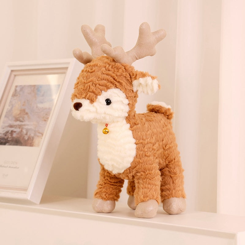 A fluffy reindeer plush with a white belly and antlers stands on a shelf beside a framed picture. It has a small bell around its neck and cream-colored accents.