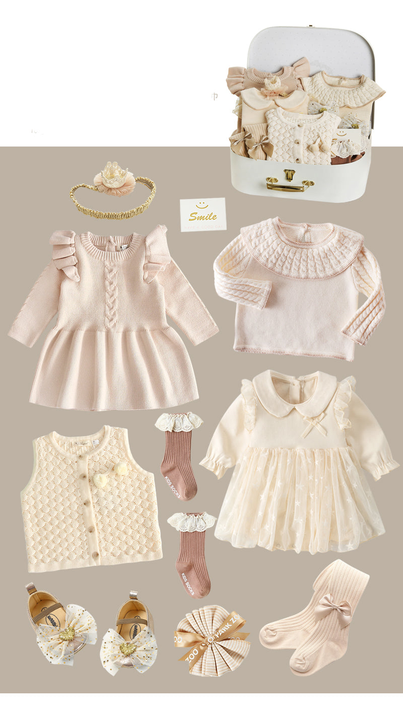 A collection of pastel baby clothes including dresses, a cardigan, socks, and shoes with bows, displayed alongside a small suitcase and a decorative headband.