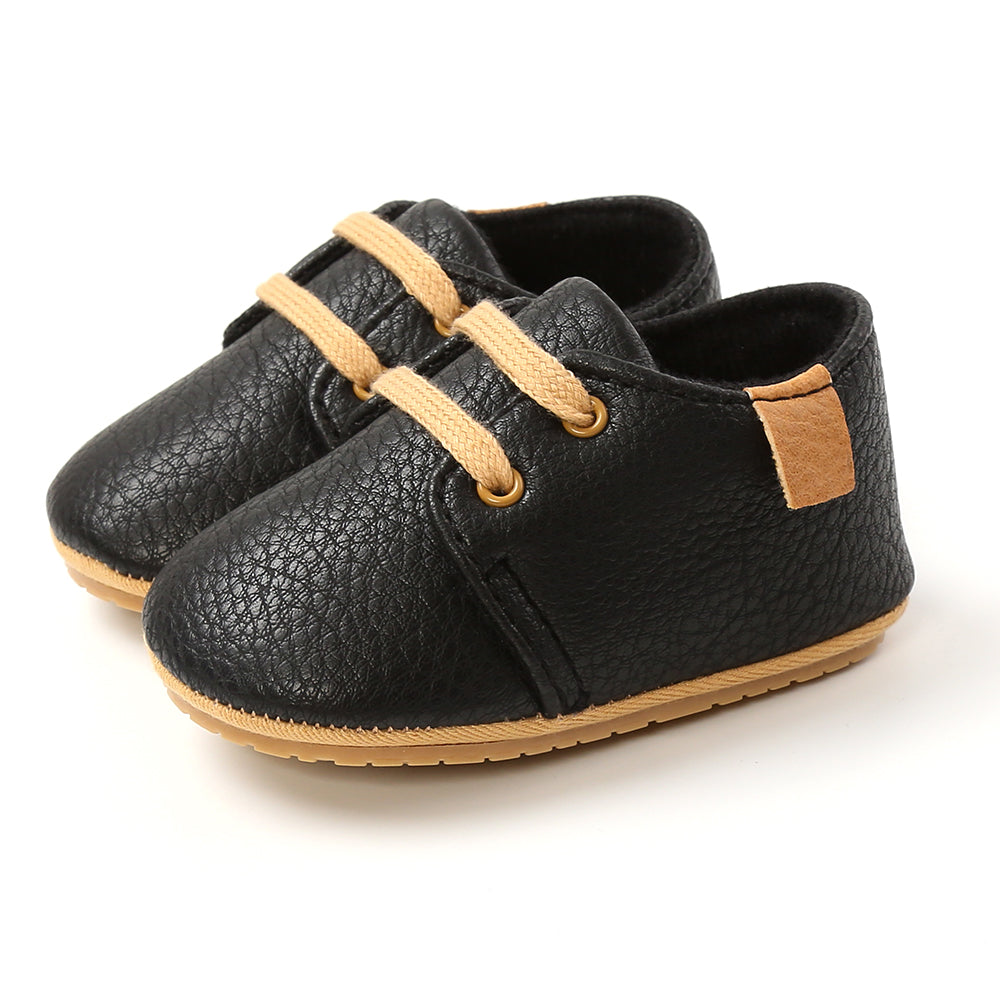 Black baby shoes with a textured leather-like finish, tan laces, and tan patches on the heels. The soles are light brown, providing a stylish and sturdy appearance.