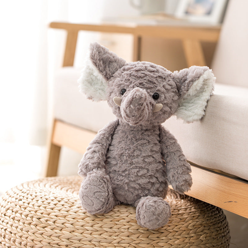 A plush gray elephant toy with large ears sits on a woven ottoman beside a beige couch in a softly lit room.