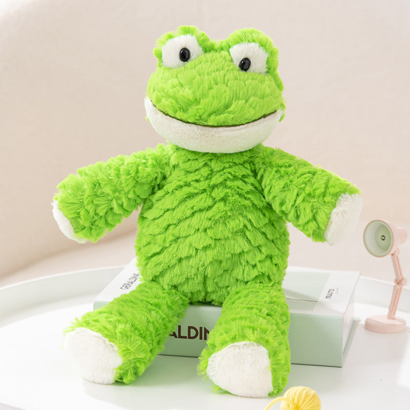 Soft green plush frog toy with big eyes and a wide smile, sitting upright on a white surface. A small lamp and a ball of yarn are in the background.