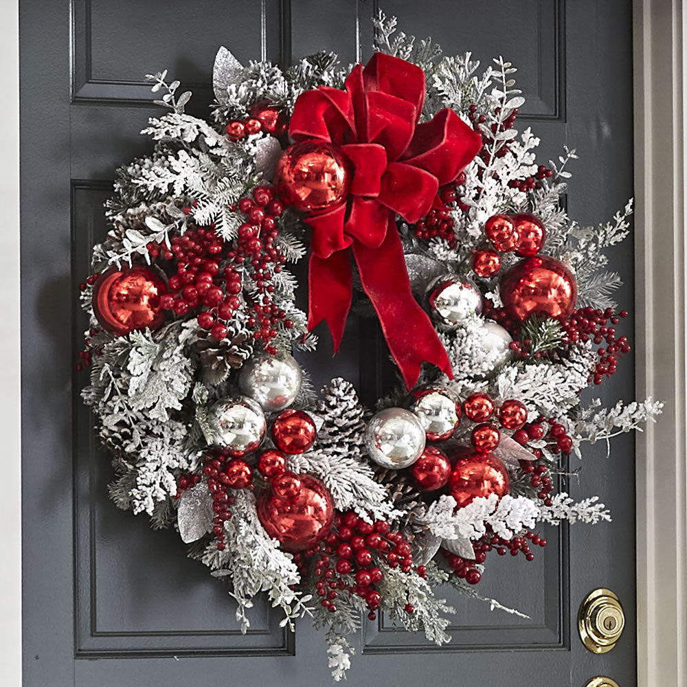 Festive wreath on a black door, adorned with red and silver ornaments, frosted greenery, and a large red velvet bow. Berries and pinecones add a traditional holiday touch.